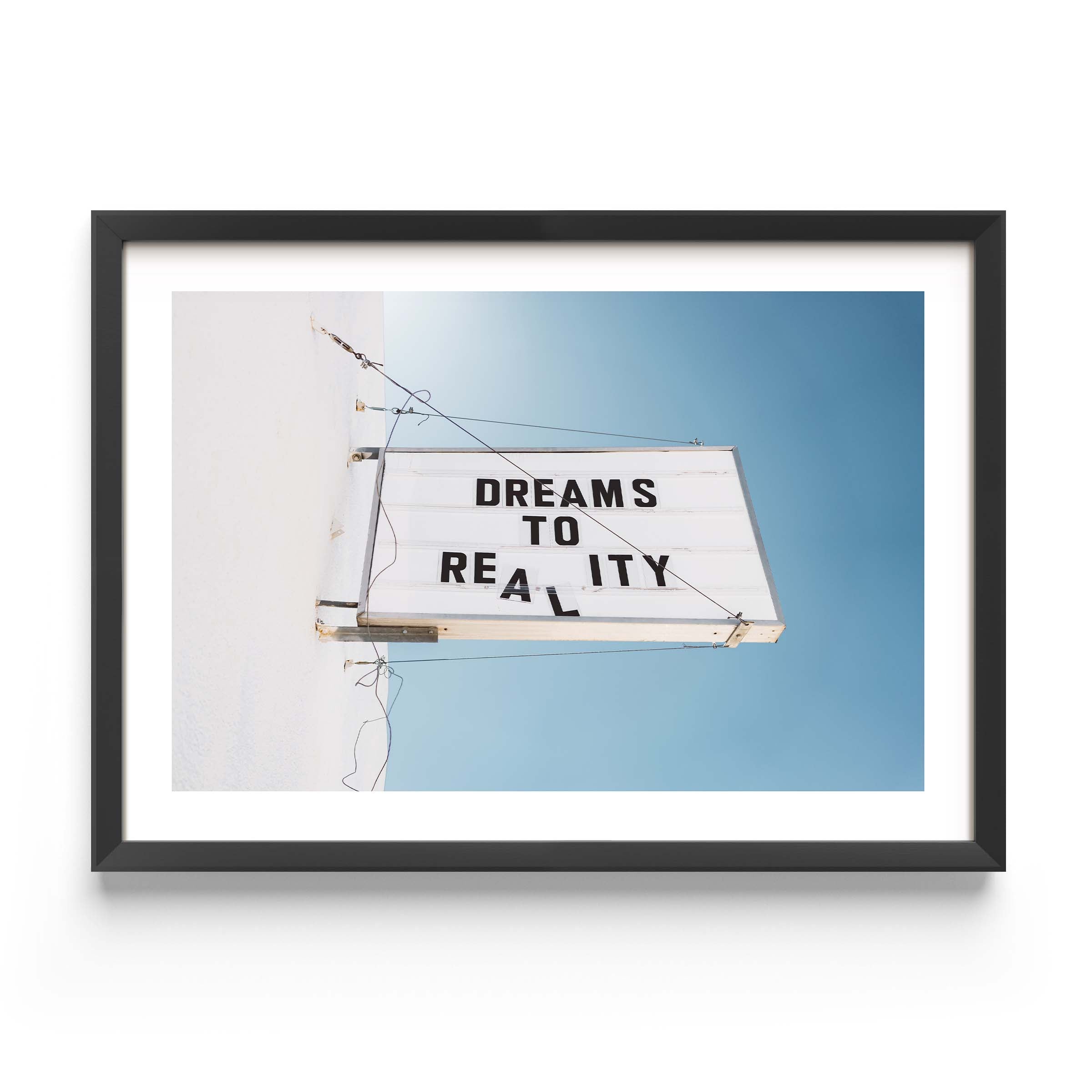 Minimal photo print of a marquee sign on a white wall reading “DREAMS TO REALITY” against a clear blue sky.