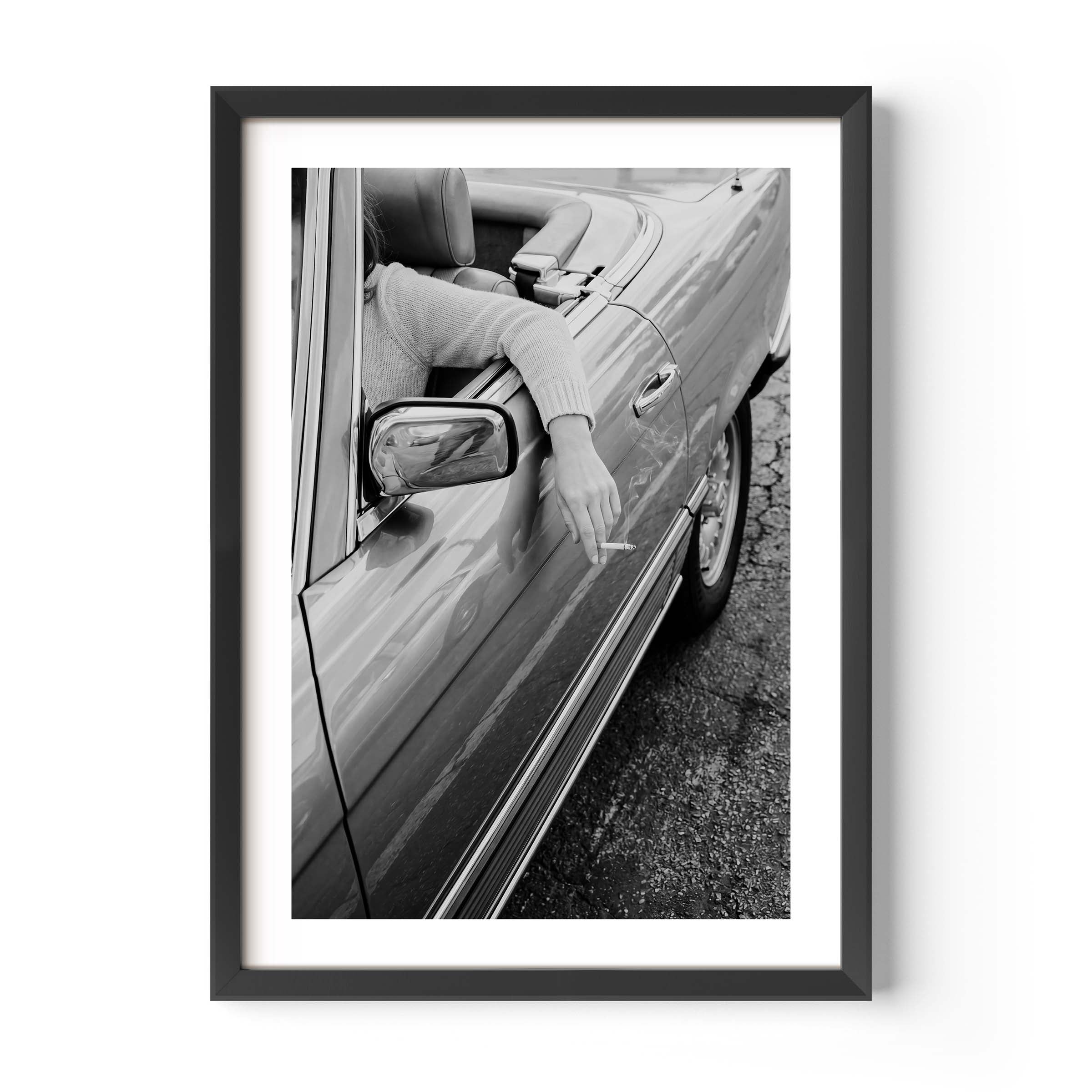 A framed black and white photographic print showing a person leaning out of a car window and smoking a cigarette, displayed against a white wall.
