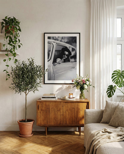 Black and white framed wall art print showing a woman in sunglasses sitting in a vintage car while applying lipstick.