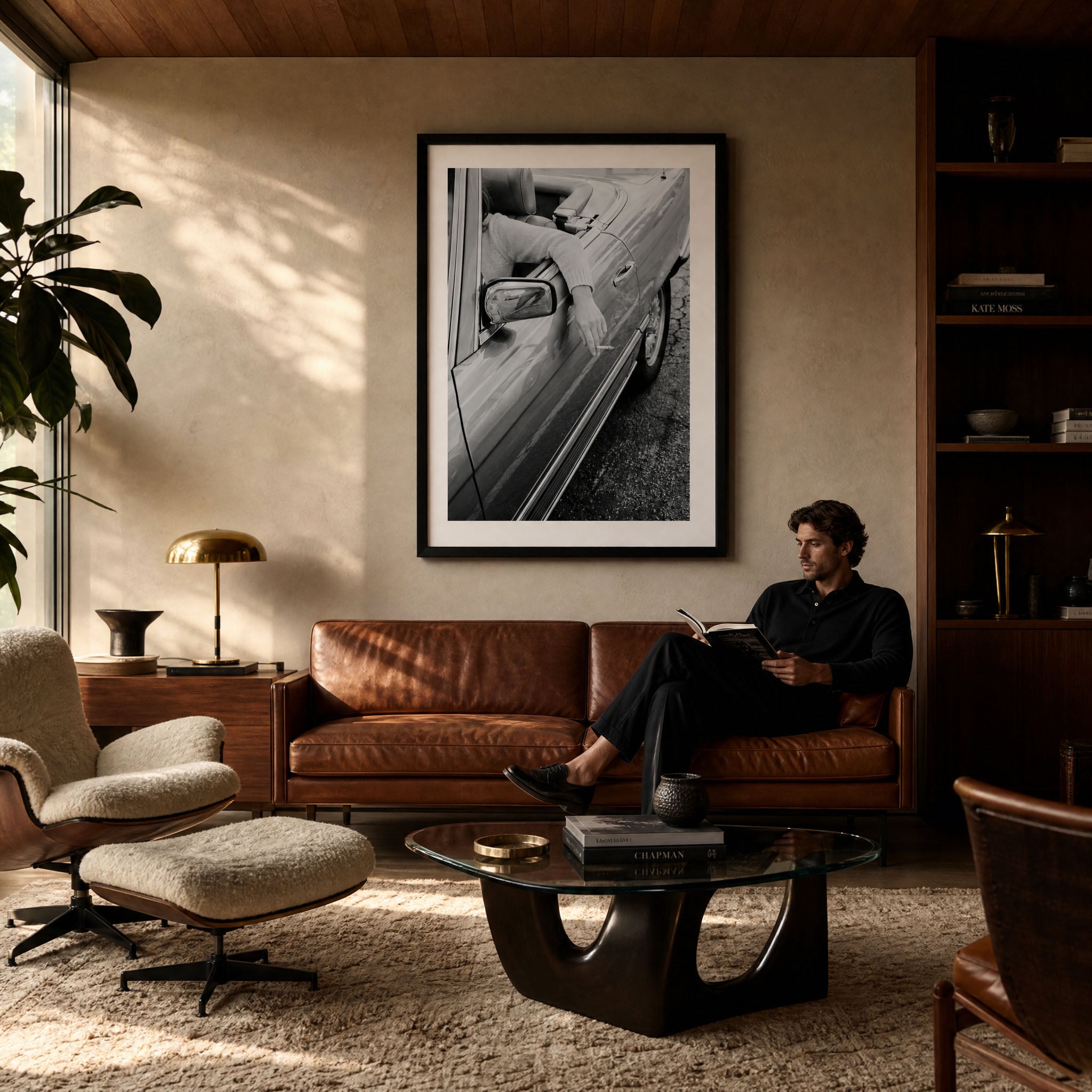 A large black and white framed art print of a vintage convertible car hangs on a beige wall in a mid-century modern living room. A man in a black shirt sits on a brown leather sofa reading a book, with a glass coffee table, leather chair, and bookshelves in the background.