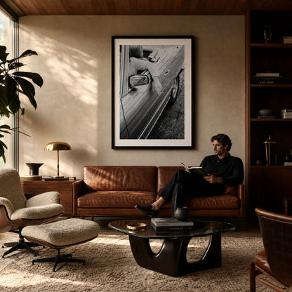 A large black and white framed art print of a vintage convertible car hangs on a beige wall in a mid-century modern living room. A man in a black shirt sits on a brown leather sofa reading a book, with a glass coffee table, leather chair, and bookshelves in the background.