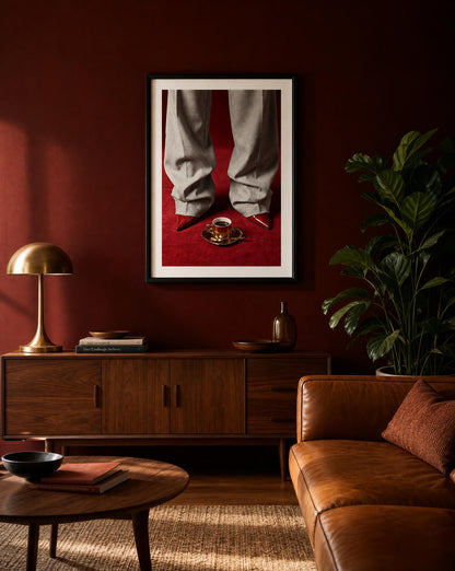 Fashion editorial wall art print showing grey wide-leg trousers, red pointed-toe stiletto heels and a gold espresso cup on a deep red velvet floor, in a black frame with white mat border.
