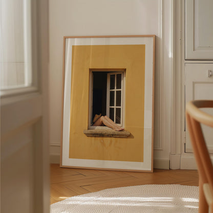 
Art print showing bare legs and feet resting on the stone sill of an open white-framed window set into a warm golden-yellow Mediterranean wall, in a natural wood frame with white mat border.