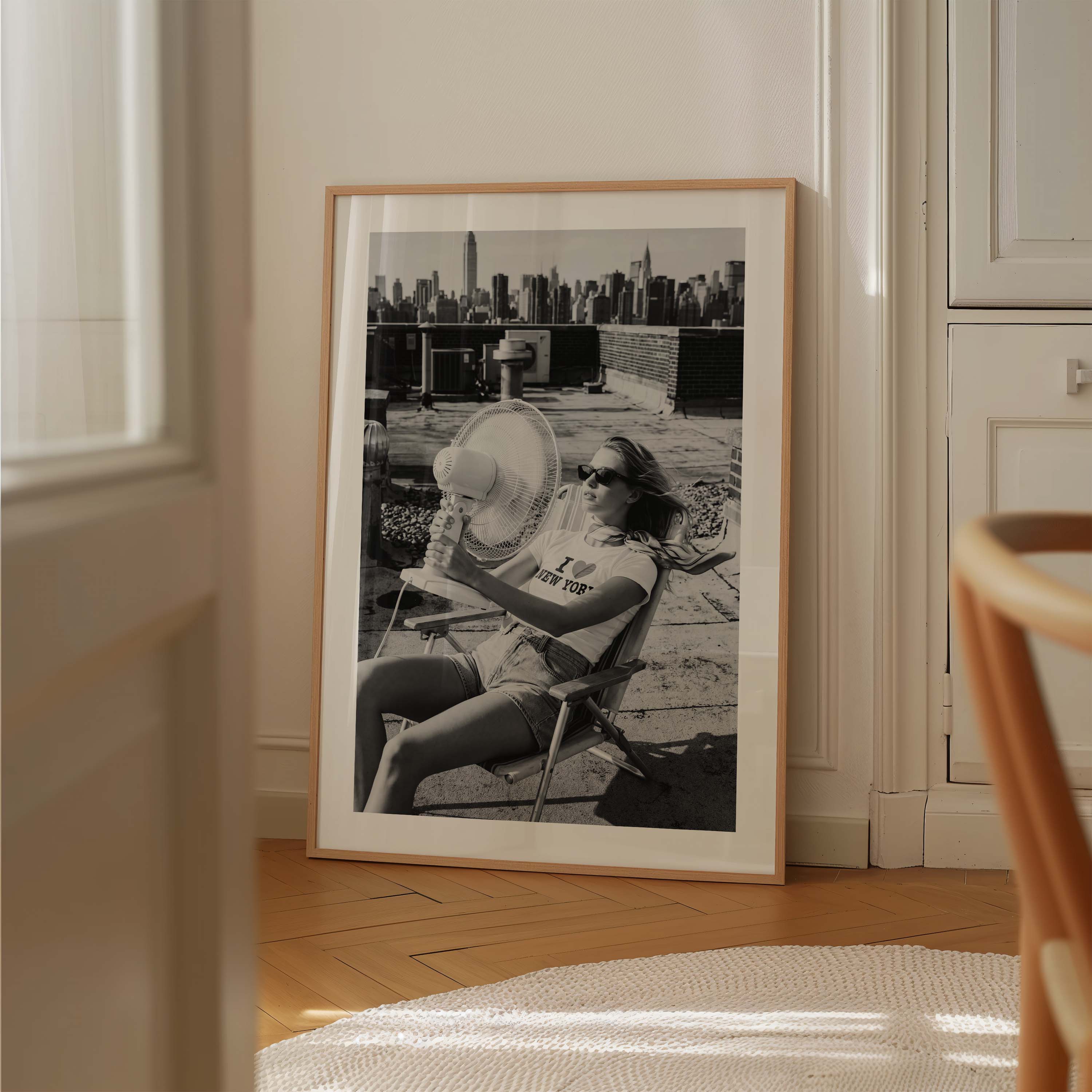 A framed black and white art print leaning against a cream-colored wall on a wooden floor. The print features a vintage photograph of a woman in a chair with a city skyline in the background, enclosed in a light wood frame.