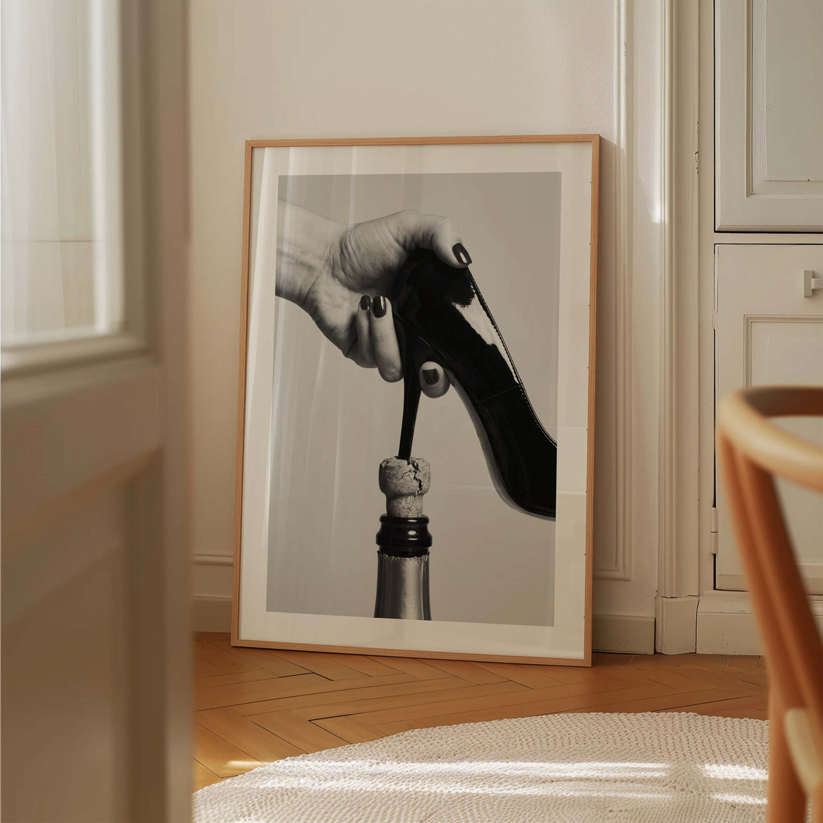 A framed black and white art print leaning against a cream-colored wall on a wooden floor, showing a hand holding a black high-heeled shoe above a bottle stopper.