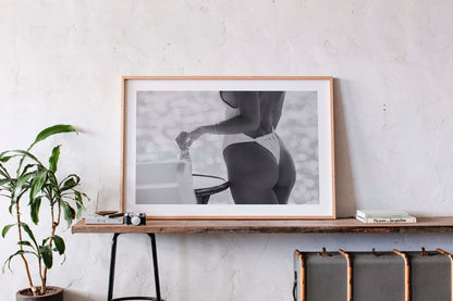 Black-and-white photo of a woman in a white one-piece holding a glass bottle by a sunlit seaside table