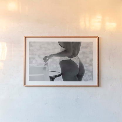 Black-and-white photo of a woman in a white one-piece holding a glass bottle by a sunlit seaside table