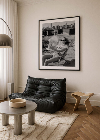 A large black and white framed art print hangs on a beige wall in a modern living room setting, featuring a black leather lounge chair, a round wooden coffee table, and a light wood stool.