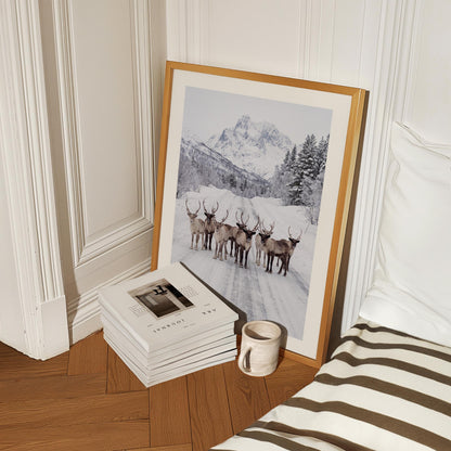 A framed black and white art print of reindeer in a snowy mountain landscape, styled in a bedroom corner next to a bed with brown and white stripes, a stack of books, and a ceramic mug.