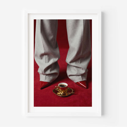 Fashion editorial wall art print showing grey wide-leg trousers, red pointed-toe stiletto heels and a gold espresso cup on a deep red velvet floor, in a black frame with white mat border.