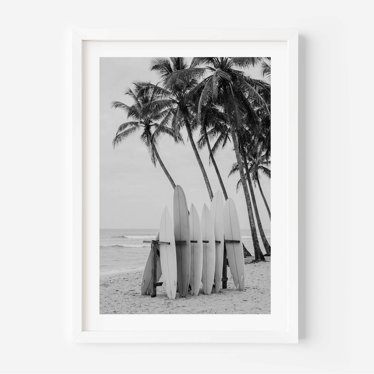 A framed black and white photographic print of surfboards leaning against a beach next to palm trees, displayed on a clean white wall.
