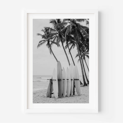 A framed black and white photographic print of surfboards leaning against a beach next to palm trees, displayed on a clean white wall.
