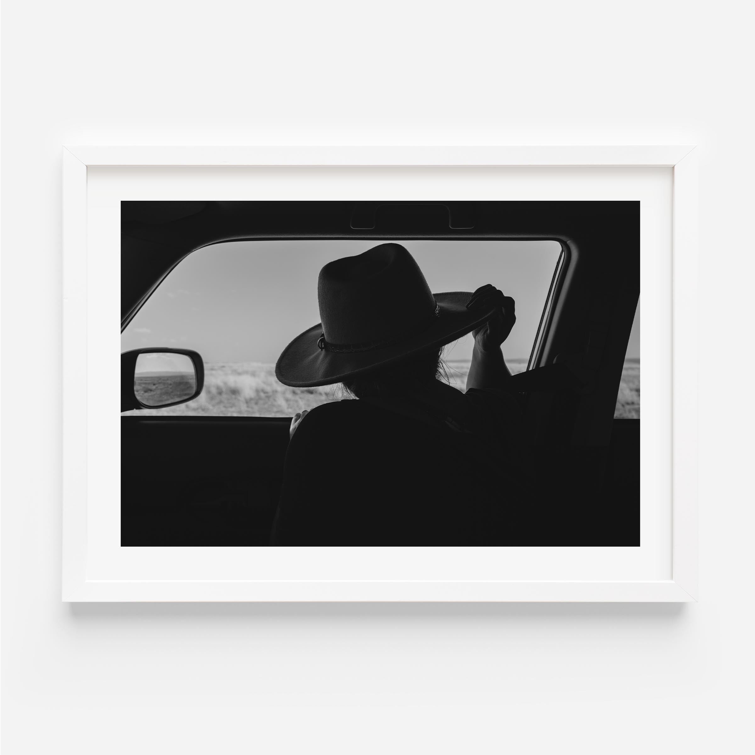 Black-and-white fine art print of a person wearing a cowboy hat seen from inside a car, looking out over an open landscape.