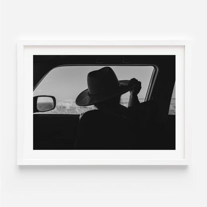 Black-and-white fine art print of a person wearing a cowboy hat seen from inside a car, looking out over an open landscape.