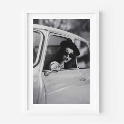 Black and white framed wall art print showing a woman in sunglasses sitting in a vintage car while applying lipstick.
