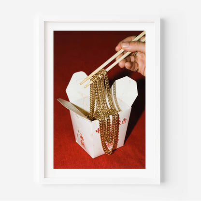 Conceptual photography print of a hand lifting gold chains with chopsticks from a Chinese takeout box on a red background, framed in black