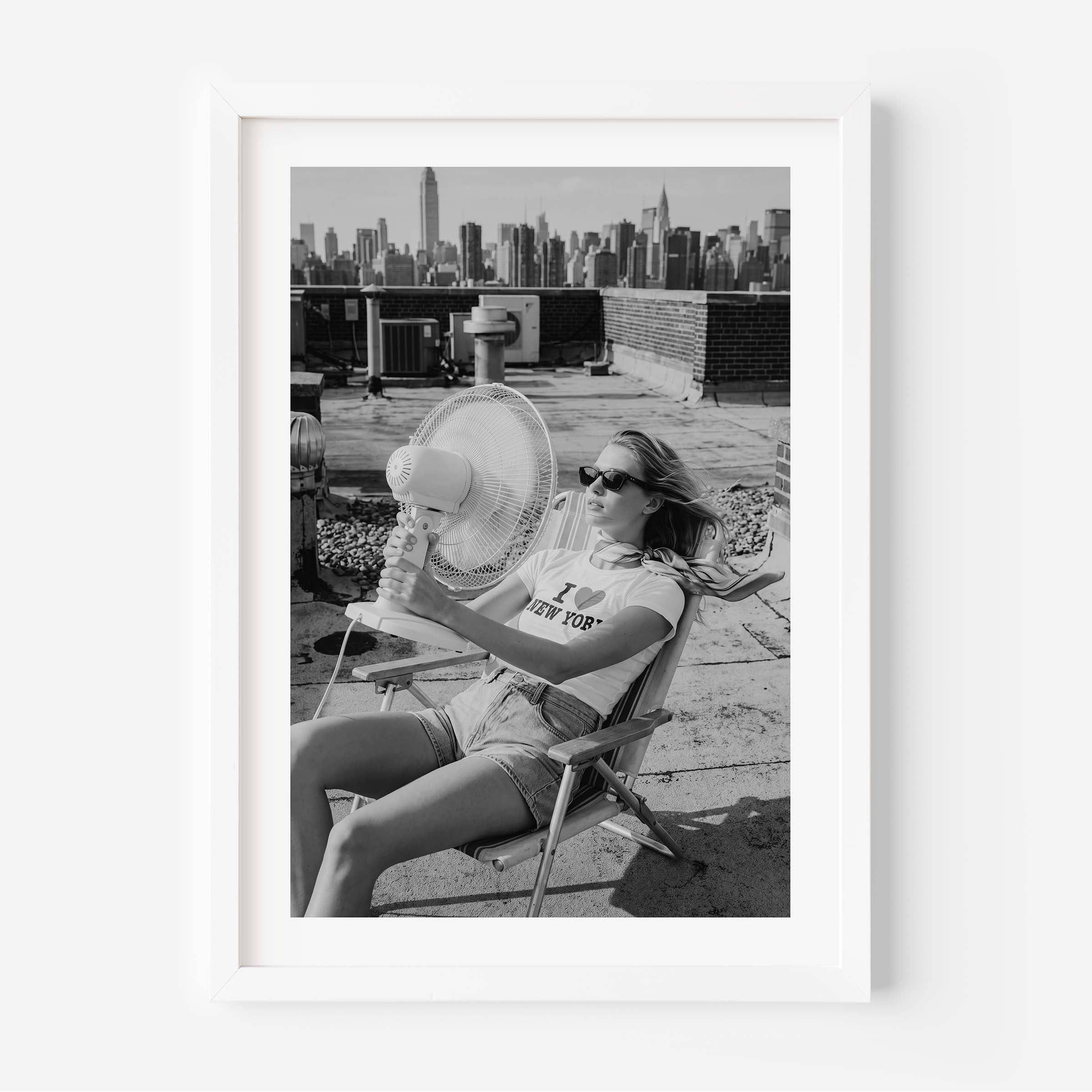A framed black and white art print featuring a woman sitting on a folding chair on a rooftop with a city skyline in the background, displayed against a clean white wall.