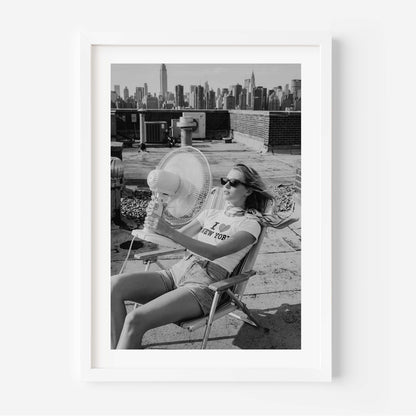 A framed black and white art print featuring a woman sitting on a folding chair on a rooftop with a city skyline in the background, displayed against a clean white wall.
