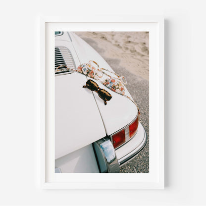 Framed wall art print showing a vintage white porsche near the beach with sunglasses and a patterned bikini placed on the back of the car.