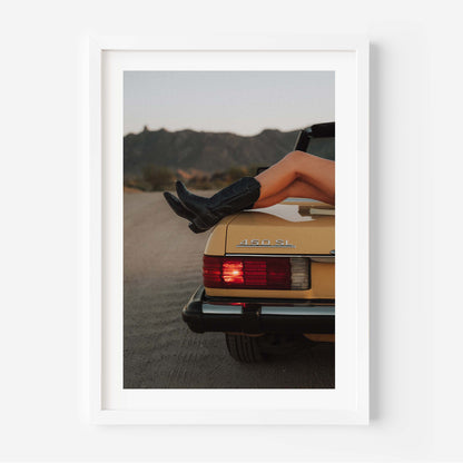 Fine art photo of the rear of a classic Mercedes 450 SL at dusk, with a person’s legs in black boots resting on the trunk and mountains in the background.