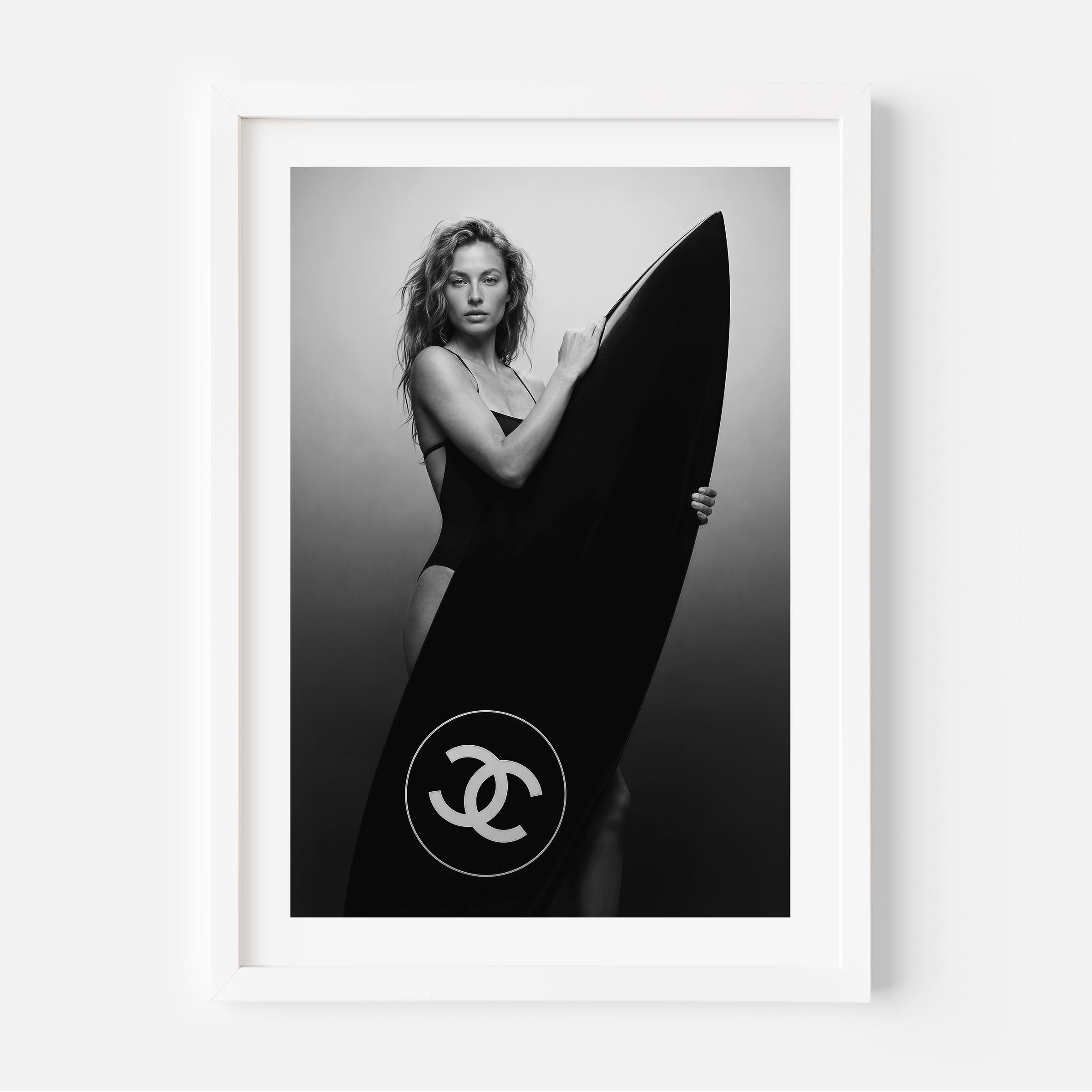 Black-and-white fine art photo of a woman in a swimsuit holding a large surfboard featuring a circular double-C style logo, against a minimal studio background.