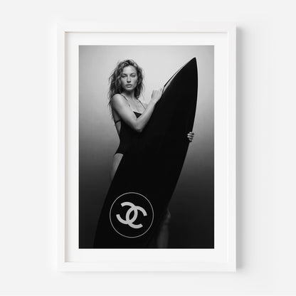 Black-and-white fine art photo of a woman in a swimsuit holding a large surfboard featuring a circular double-C style logo, against a minimal studio background.