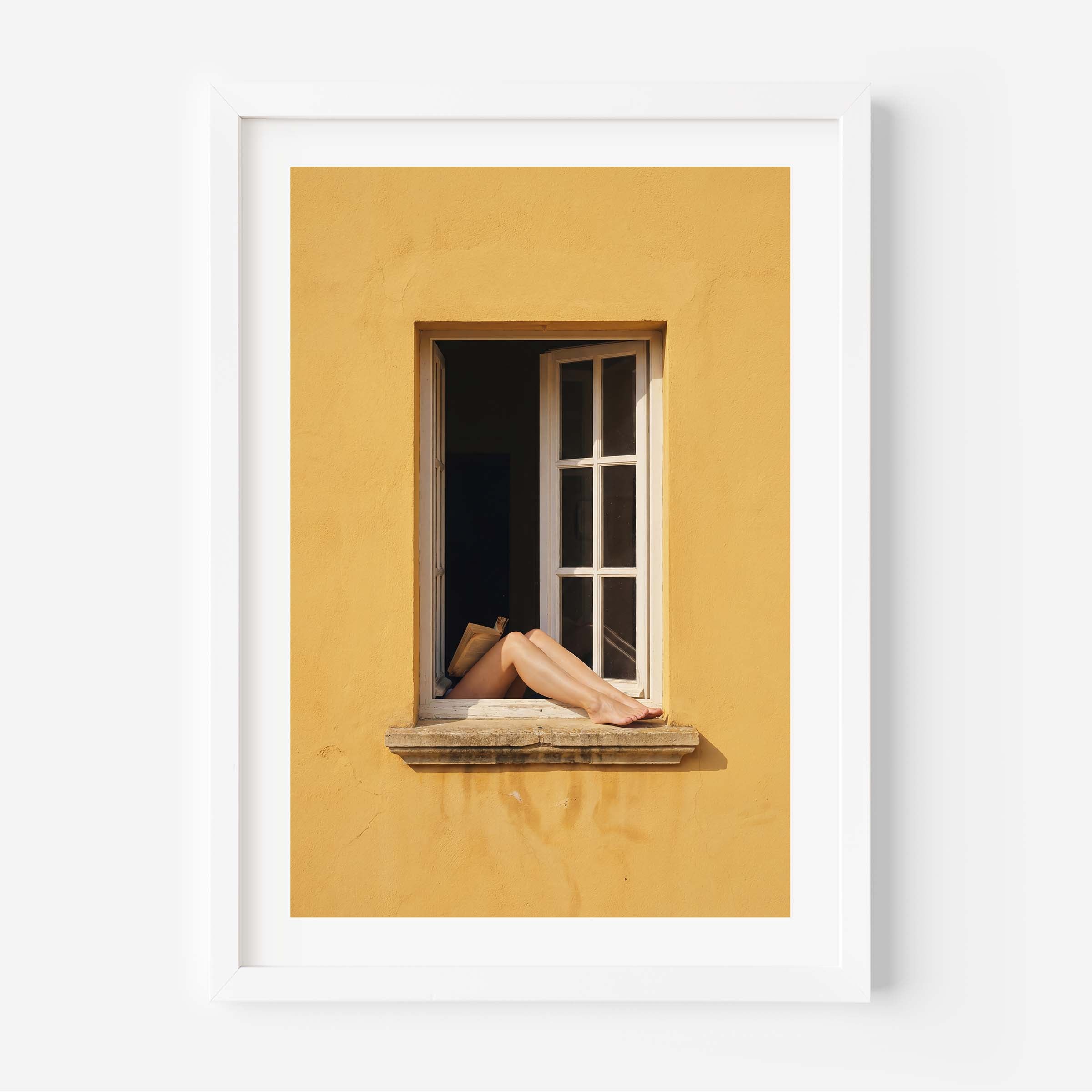 
Art print showing bare legs and feet resting on the stone sill of an open white-framed window set into a warm golden-yellow Mediterranean wall, in a natural wood frame with white mat border.