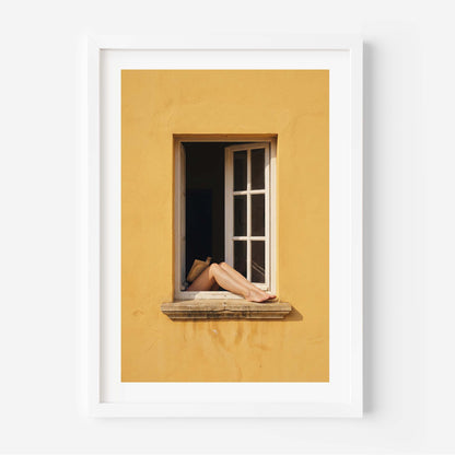 
Art print showing bare legs and feet resting on the stone sill of an open white-framed window set into a warm golden-yellow Mediterranean wall, in a natural wood frame with white mat border.