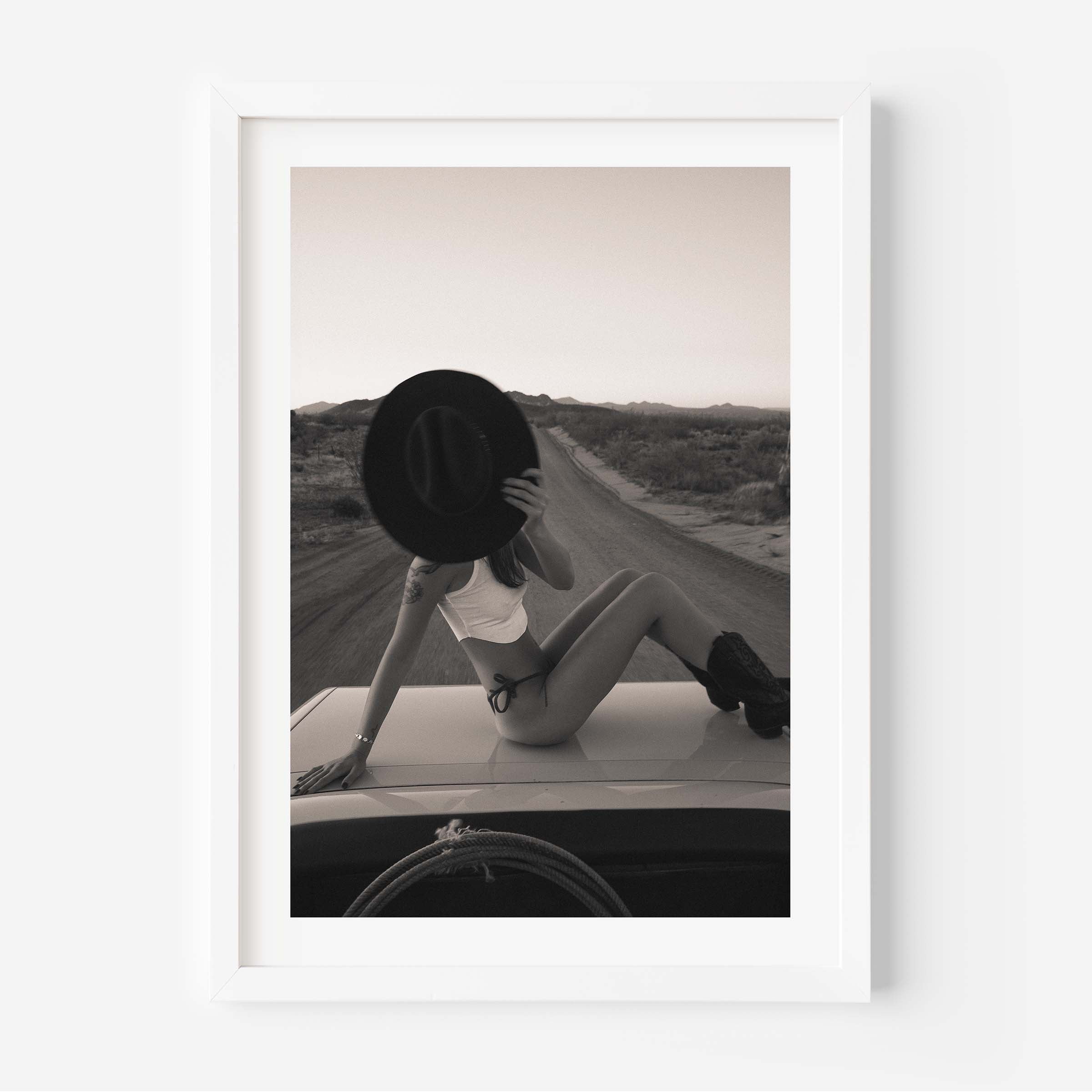 Fine art photo print of a woman sitting on a car roof on a desert road, holding a wide-brim hat in front of her face at sunset.