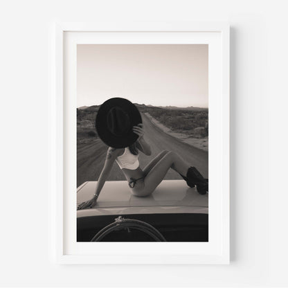 Fine art photo print of a woman sitting on a car roof on a desert road, holding a wide-brim hat in front of her face at sunset.