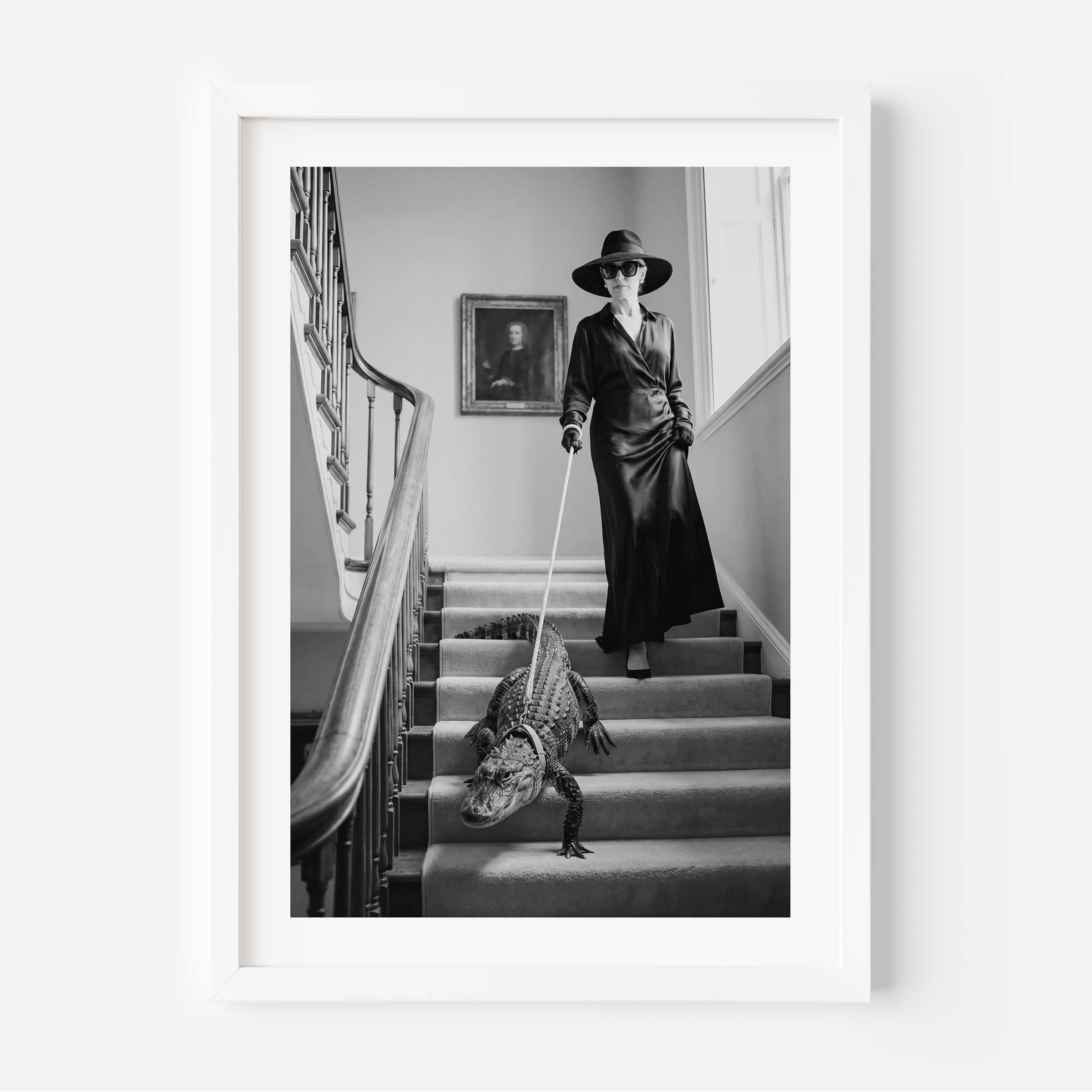 A framed black and white art print depicting a woman in a long coat and wide-brimmed hat walking a large alligator down a grand staircase, shown against a clean white background.