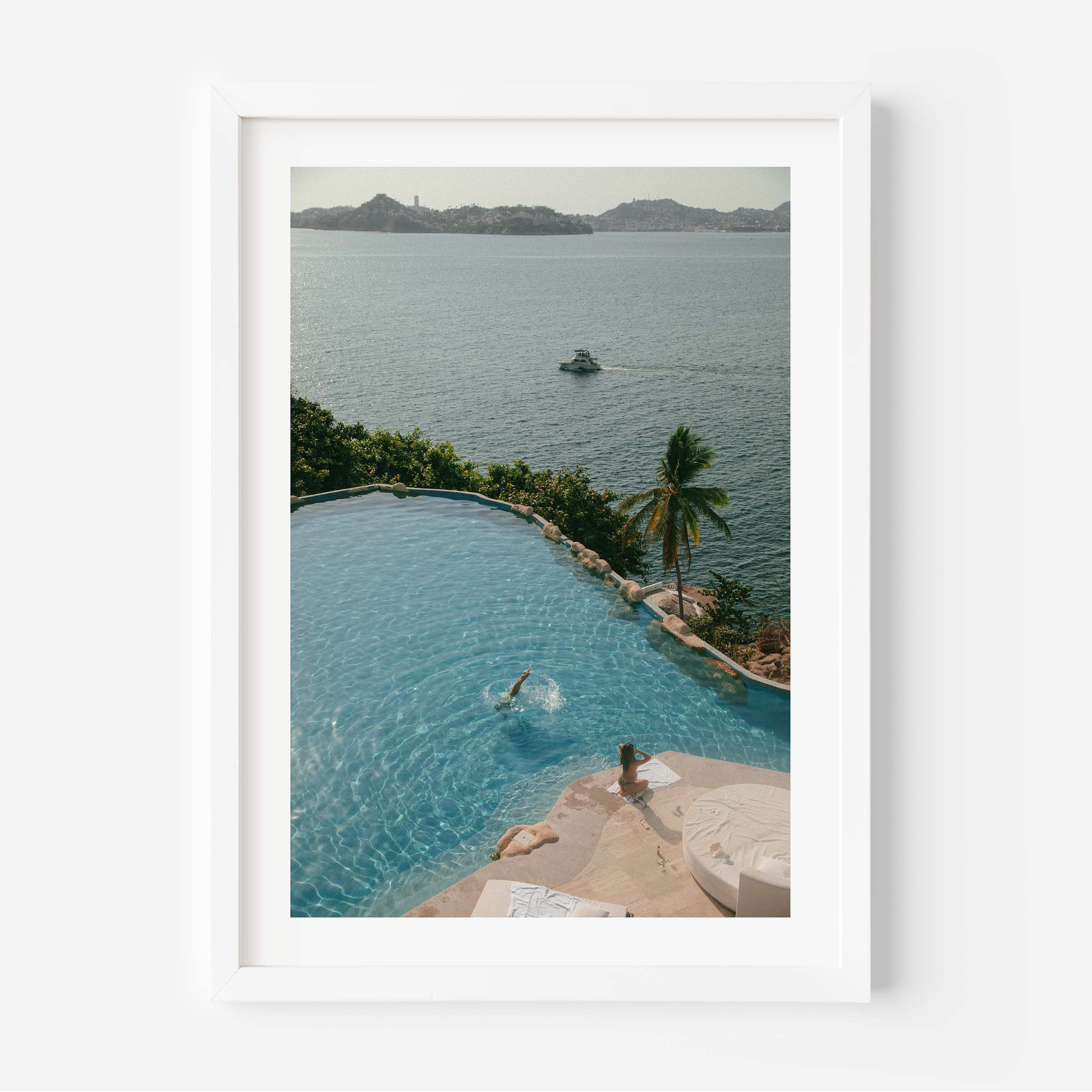 Framed color photograph of a diver in a turquoise infinity pool above a tropical bay with palm tree, sunlounger and boat in the distance — by Britt Van der Meyden.