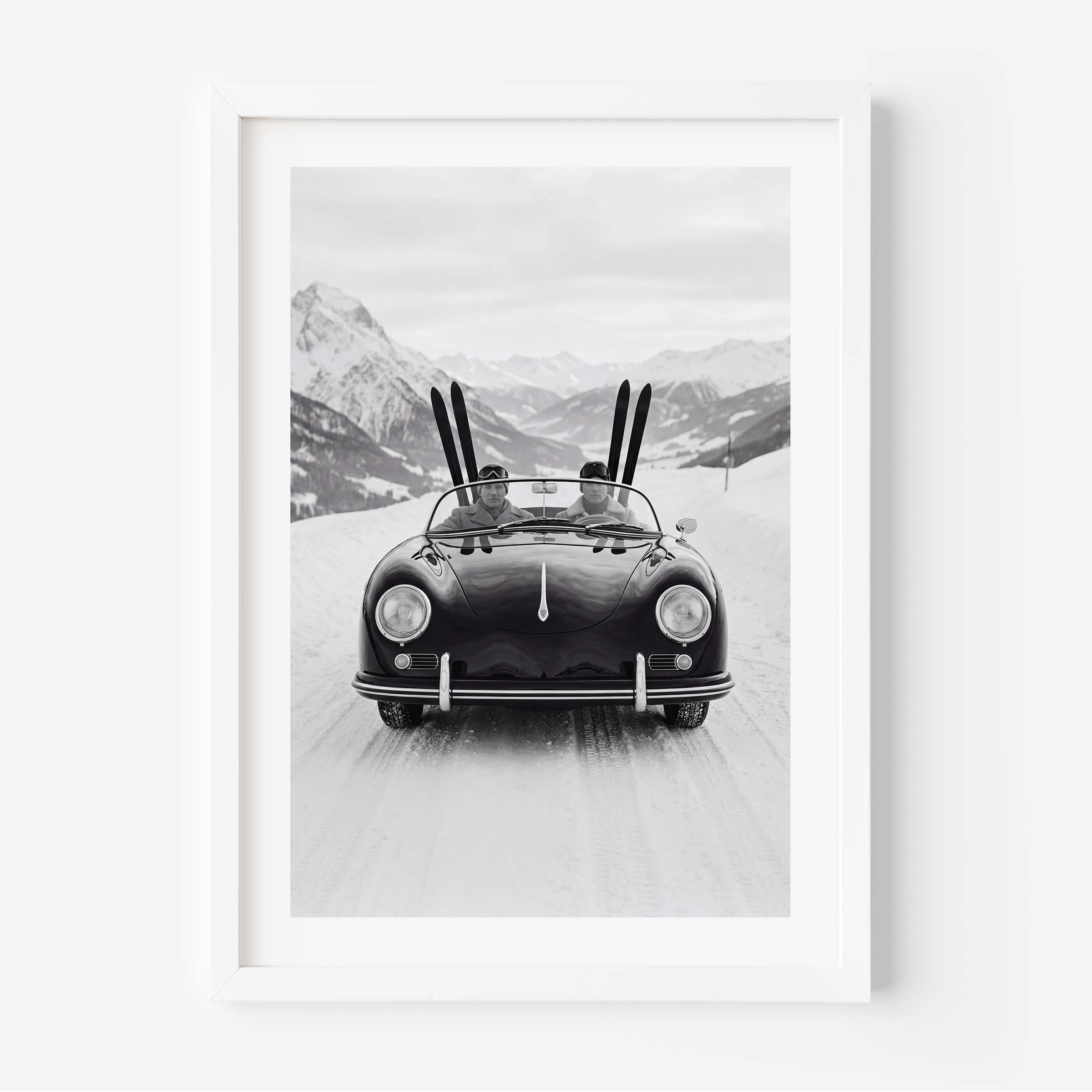 Black-and-white photo of a vintage convertible driving on a snowy mountain road, with two people inside and skis standing upright behind them, alpine peaks in the background.