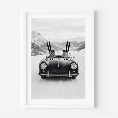 Black-and-white photo of a vintage convertible driving on a snowy mountain road, with two people inside and skis standing upright behind them, alpine peaks in the background.