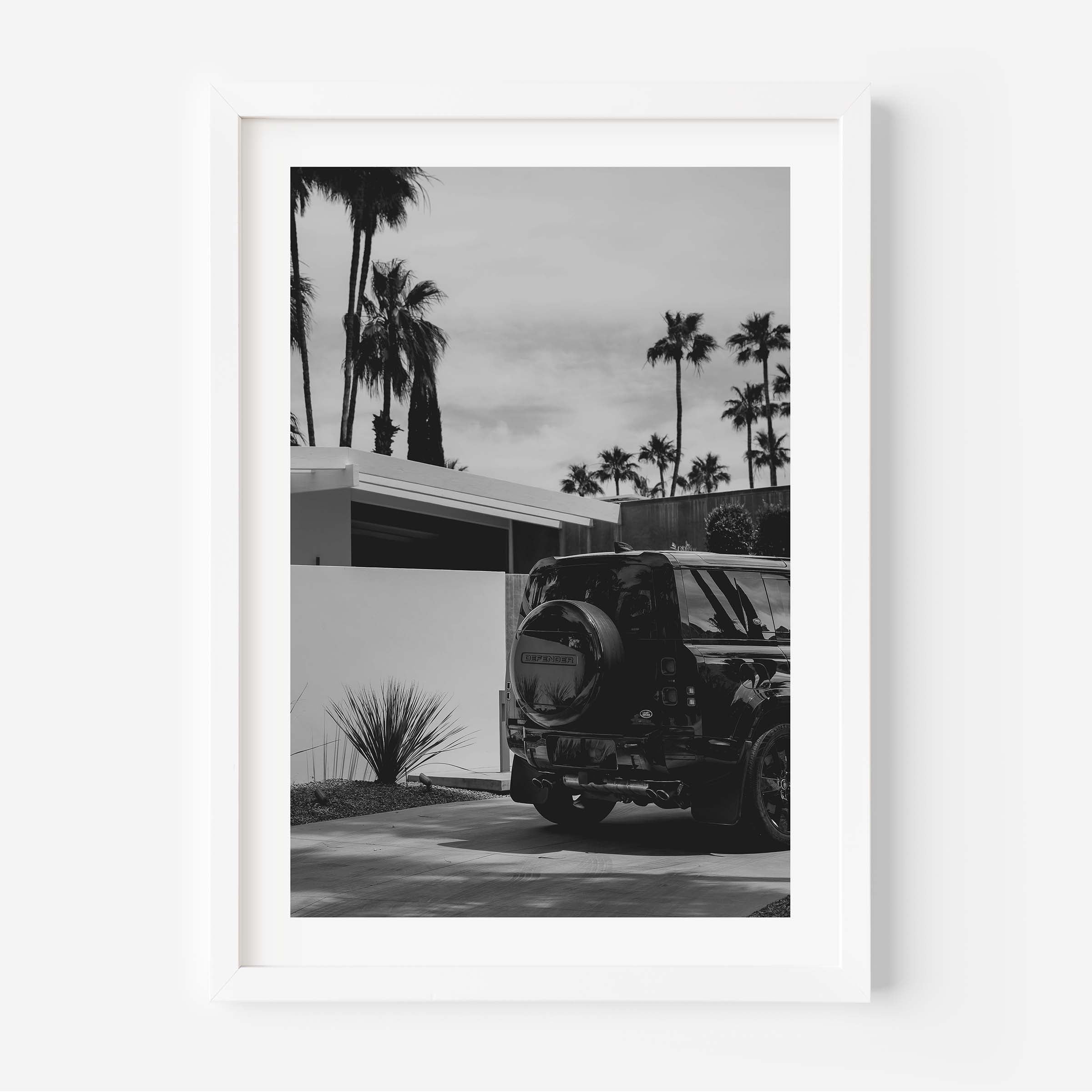 Black-and-white photo of a Land Rover Defender parked in a driveway beside a modern home, with tall palm trees and a bright sky in the background.