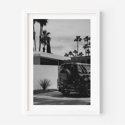 Black-and-white photo of a Land Rover Defender parked in a driveway beside a modern home, with tall palm trees and a bright sky in the background.