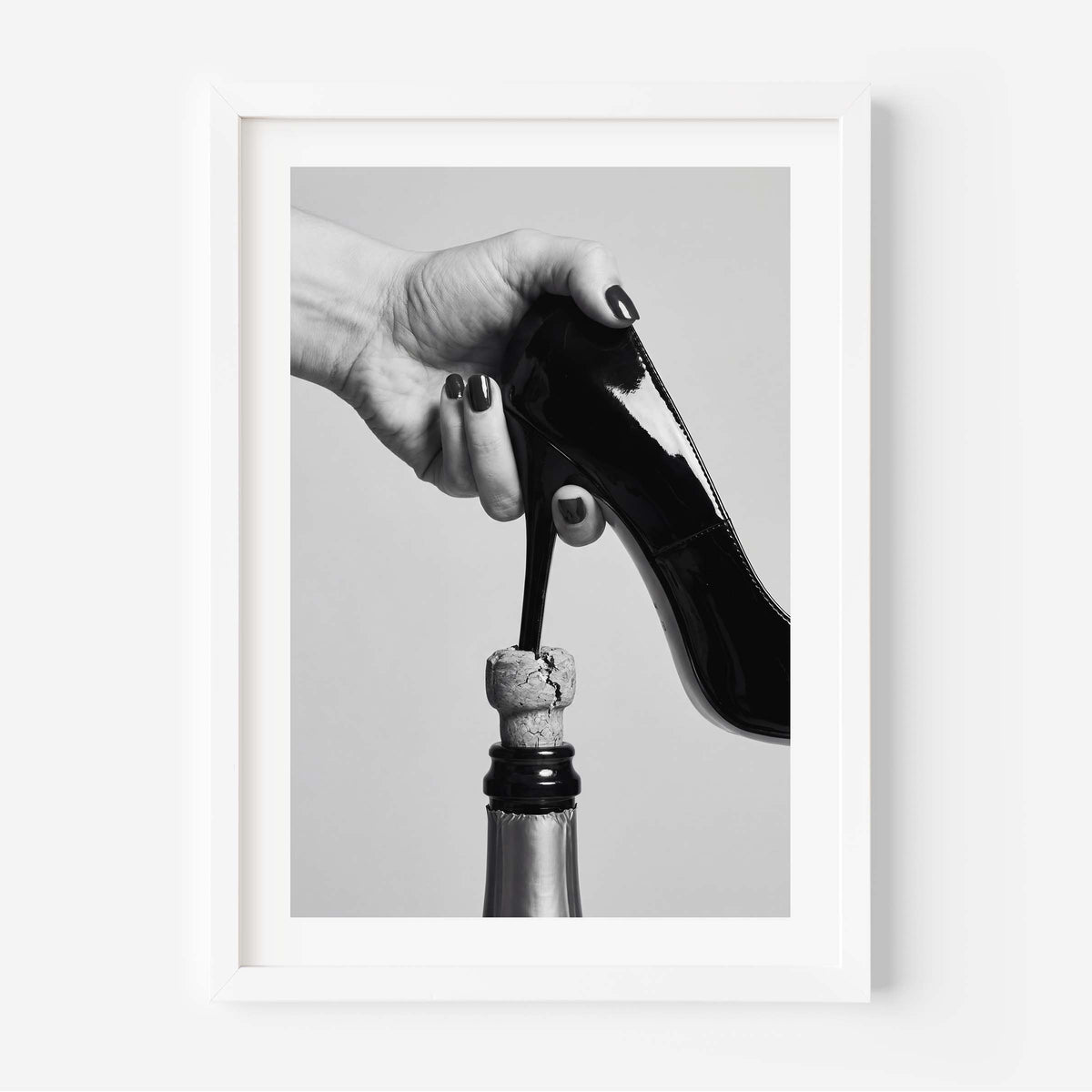 A framed black and white art print depicting a hand with dark nail polish holding a black patent leather high heel above a champagne bottle cork, set against a light gray background.