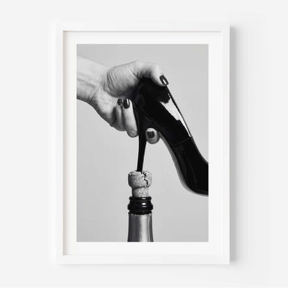 A framed black and white art print depicting a hand with dark nail polish holding a black patent leather high heel above a champagne bottle cork, set against a light gray background.