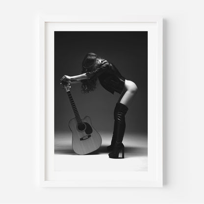 Black-and-white photo of a woman in a leather jacket and thigh-high boots leaning over an acoustic guitar in a studio, framed with white mat.