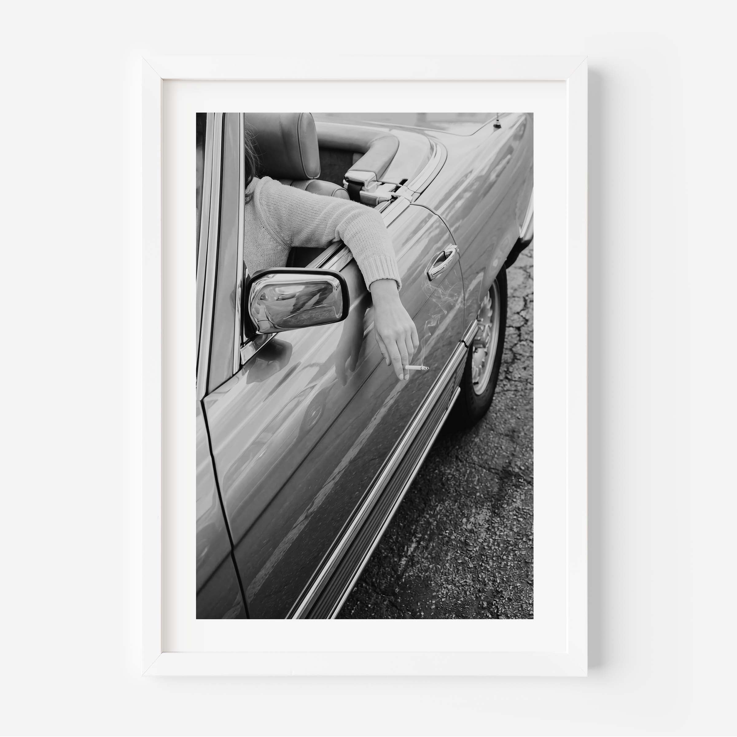 A framed black and white art print depicting a person leaning out of a convertible car and smoking a cigarette, shown in a clean white frame against a white wall.