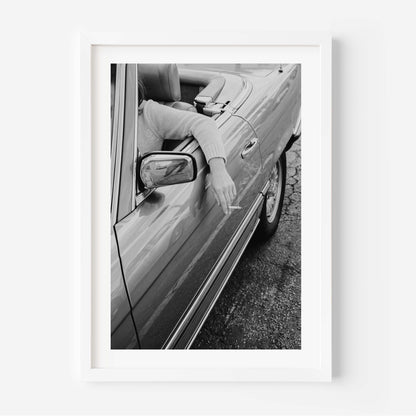 A framed black and white art print depicting a person leaning out of a convertible car and smoking a cigarette, shown in a clean white frame against a white wall.