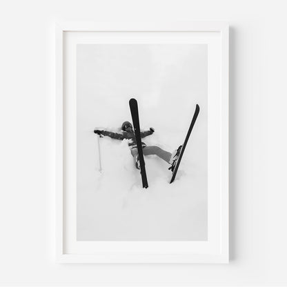 Black and white fine art print of a skier lying in deep snow with skis and pole scattered, framed in black.