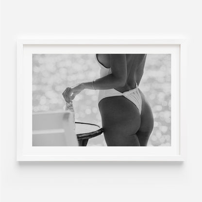 Black-and-white photo of a woman in a white one-piece holding a glass bottle by a sunlit seaside table