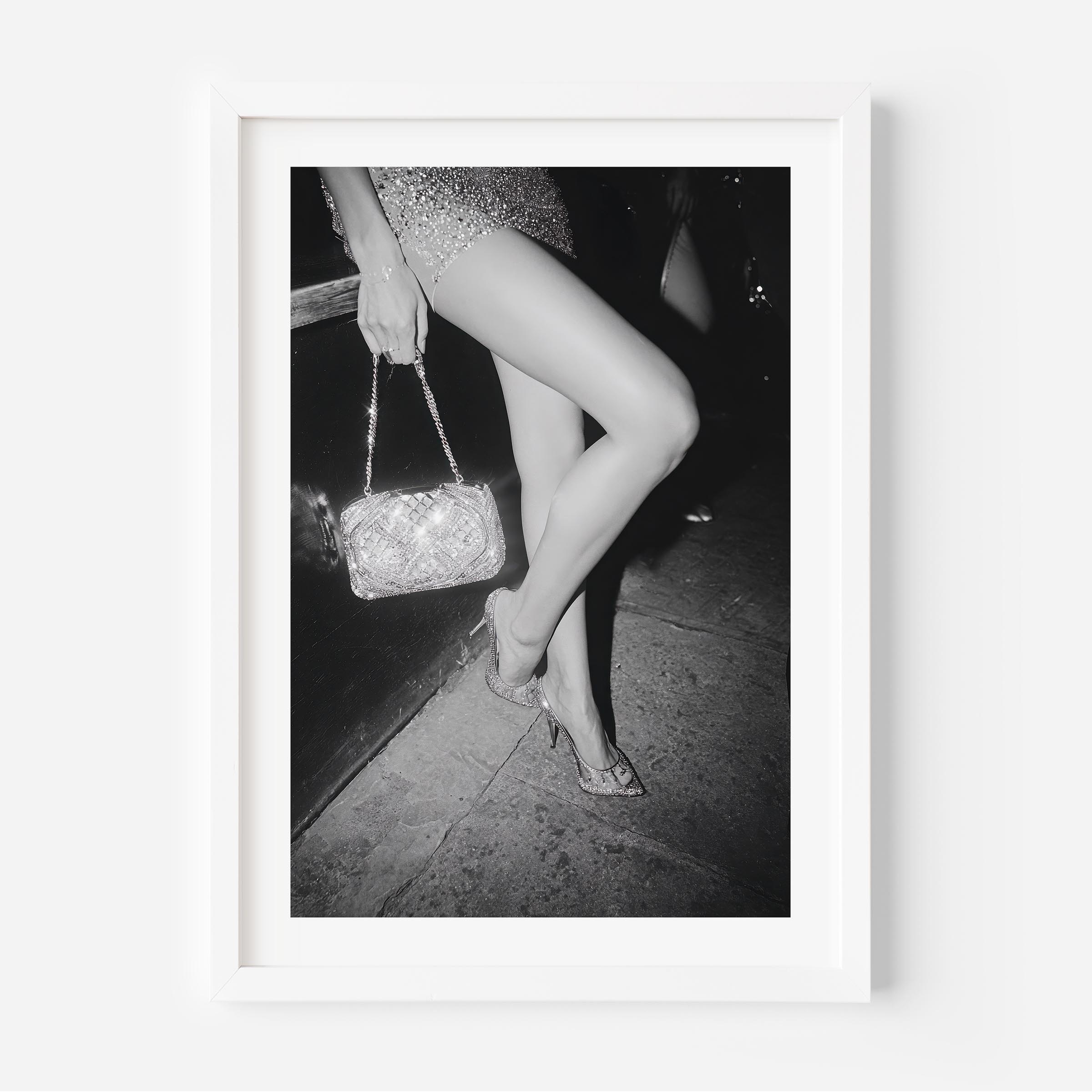 Black-and-white fine art photograph of a woman’s legs in high heels and sequin dress, holding a glittering handbag on a city sidewalk.