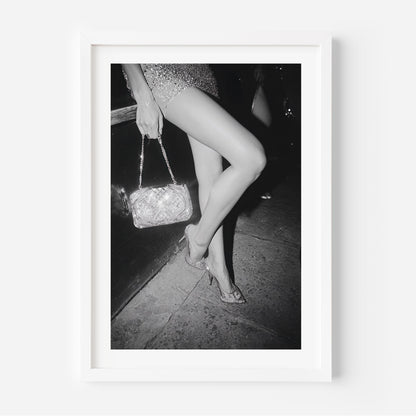 Black-and-white fine art photograph of a woman’s legs in high heels and sequin dress, holding a glittering handbag on a city sidewalk.