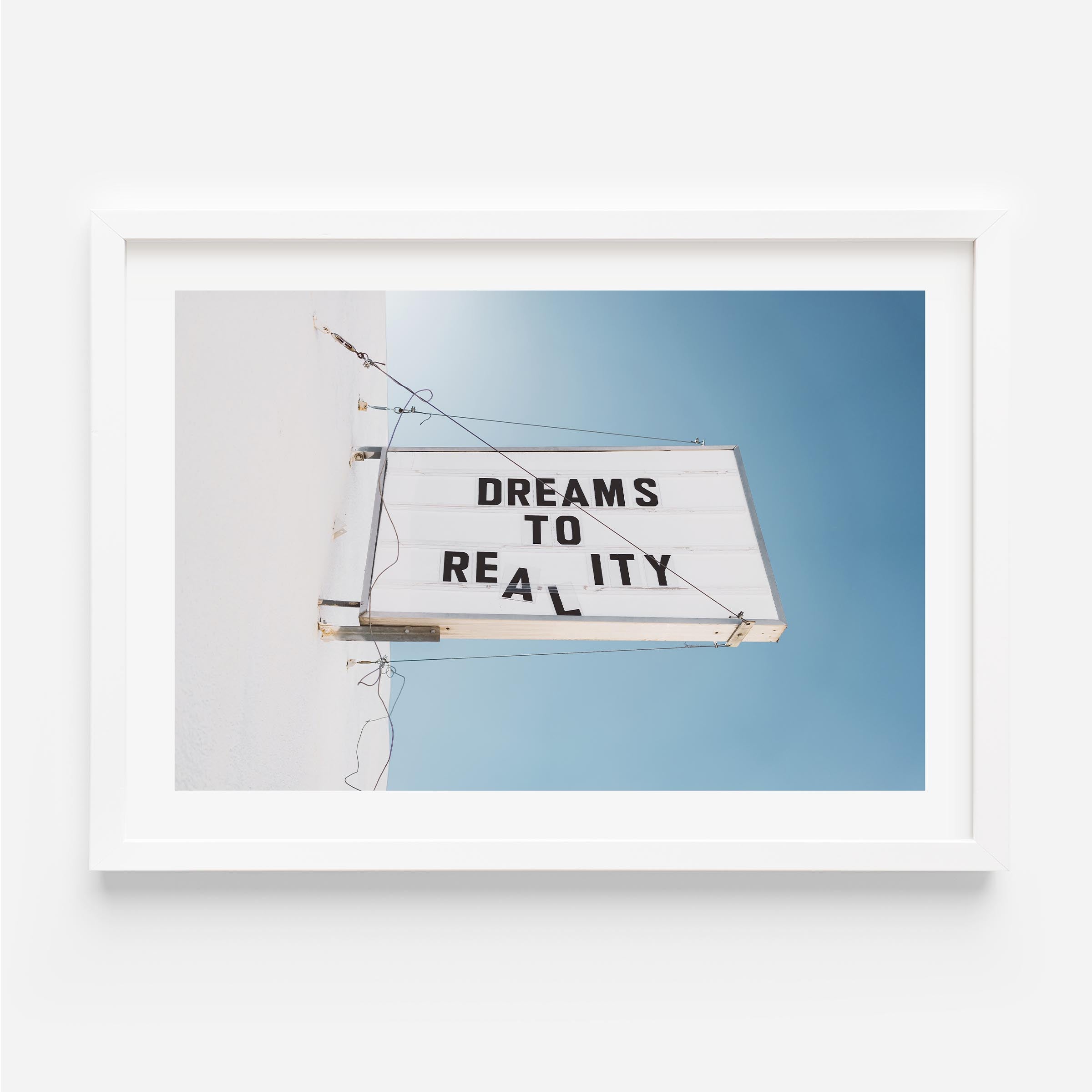 Minimal photo print of a marquee sign on a white wall reading “DREAMS TO REALITY” against a clear blue sky.