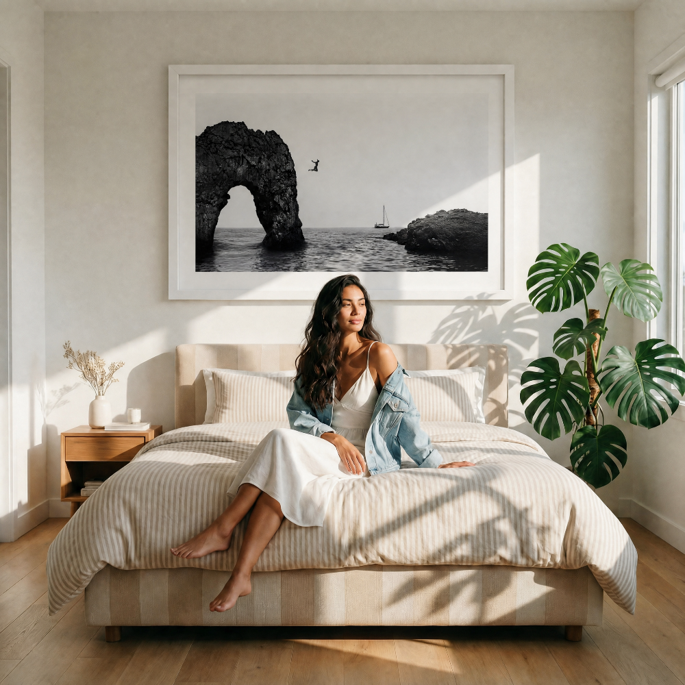 Woman sitting on a bed in a bright bedroom with a large framed photograph of a coastal scene above her.