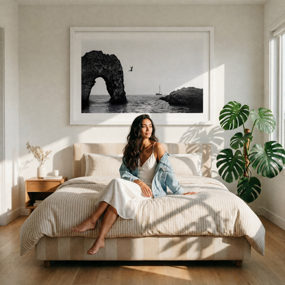 Woman sitting on a bed in a bright bedroom with a large framed photograph of a coastal scene above her.