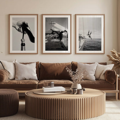 Three framed black and white art prints featuring fashion and boating imagery are displayed on a beige wall above a brown velvet sofa in a modern living room setting.