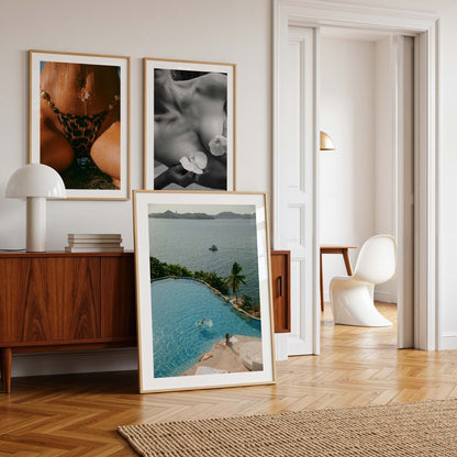 Framed color photograph of a diver in a turquoise infinity pool above a tropical bay with palm tree, sunlounger and boat in the distance — by Britt Van der Meyden.